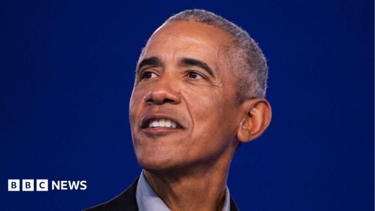 Barack Obama, standing on a stage and smiling.