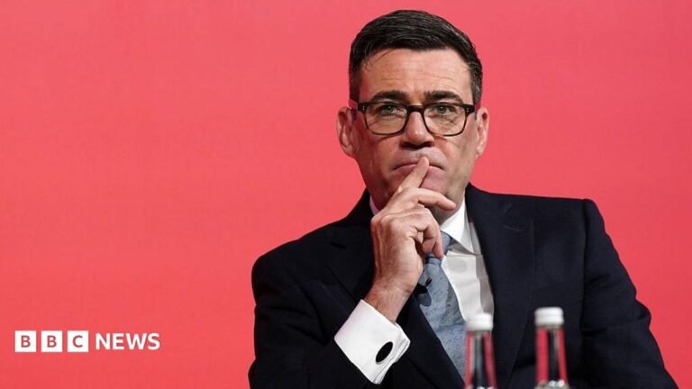 Chris Mason: Why Andy Burnham’s about-turn on Manchester City matters Andy Burnham looks pensive while sitting down with a red background.