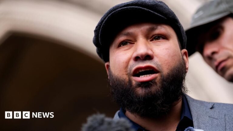 A man wearing a flat cap stands outside a court building and addresses members of the press.