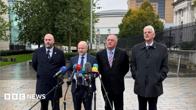 Drumcree: Legal bid to overturn march ban fails Four men stand behind six microphones. They stand on light-grey and beige bricks in front of white-stone buildings.