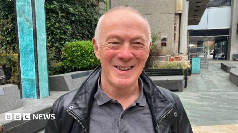 Ex-Manchester leader defends deals with Man City owners Richard Leese stands in front of stone buildings outside on a high street. He wears a grey polo shirt and black leather jacket. He has short grey hair and is smiling.