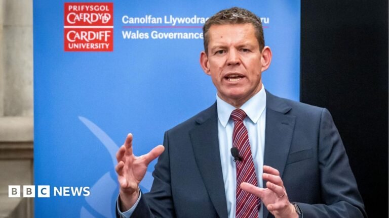 First Minister Rhun ap Iorwerth promises to look at Welsh council powers The first minister Rhun ap Iorwerth stood in front of a banner advertising Cardiff University's Welsh governance centre. The banner is blue and has a red Cardiff University logo. Rhun wears a grey suit, white shirt, and red tie with diagonal stripes.