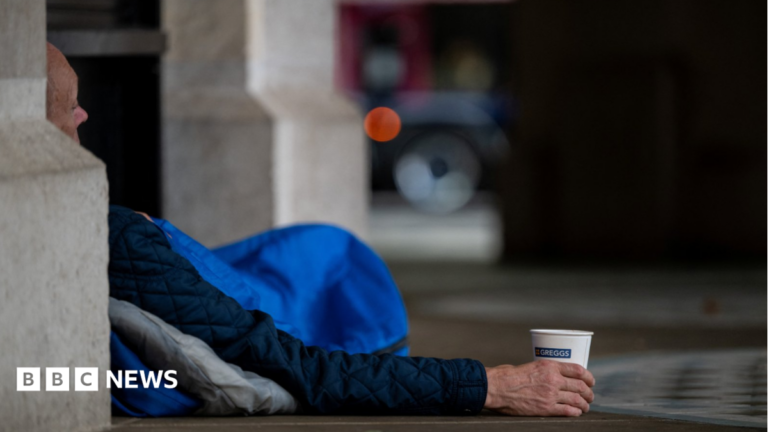 Funding set to double for plan to get rough sleepers off streets by Christmas A homeless person lies in a London doorway in a blue sleeping bag. Part of the man's face is visible around the corner of a building and he is holding a paper cup.