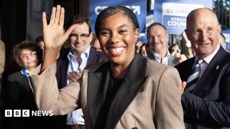 Kemi Badenoch - dressed in a light brown winter coat - waves at the camera as she arrives at the Tory conference, with applauding supporters