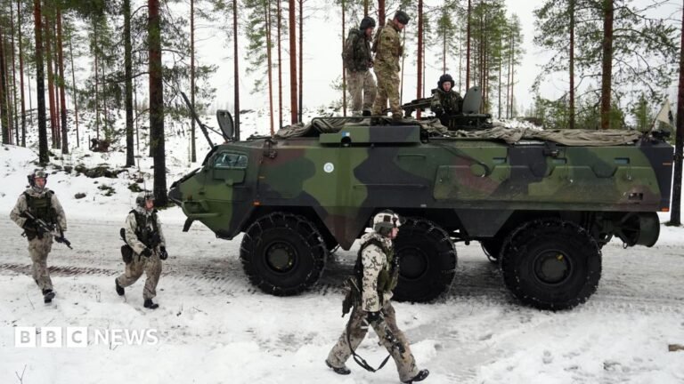John Healey tells departments to make 1% cuts to fund defence Three soldiers walk alongside an armoured tank in a snowy landscape. Three soldiers sit on top of the tank.