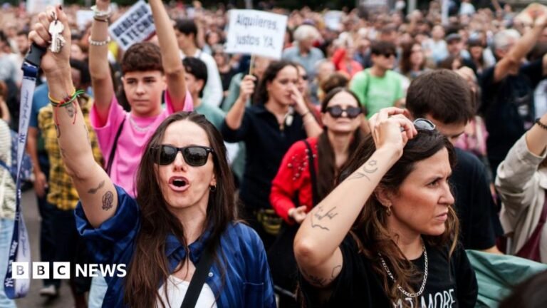 A large crowd of people marching through the streets of Valencia. Many are holding signs with a large one in the centre of the image reading 'els inversors son els invasors'.