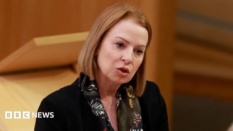 Reform MSP Amanda Bland resigns to sit as independent A woman with shoulder-length fair hair in a close-up. She is wearing a black jacket and floral scarf, with a wooden podium in the background
