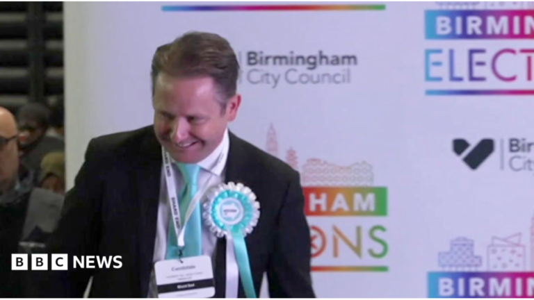 Alan Feeney on the stage after being elected a councillor at election in May 2026. He is wearing a dark jacket and white shirt with a light blue Reform coloured tie and a similar coloured rosette. There is election branding behind him.