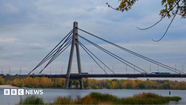 A view of the Pivnichnyi Bridge in Kyiv, Ukraine. There are cars driving across it and water running below with orange and green trees lining the banks.