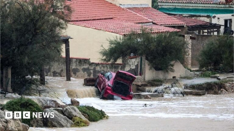 A woman wearing shorts and a white t-shirt wades through brown water in front of her house