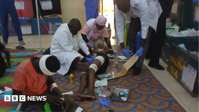 Sudan army drones hit mourners and fuel market killing more than 300 people, sources say An man is wearing a green helmet with chin straps. He is wearing camouflage clothing and looking up into the distance. He is surrounded by trees.