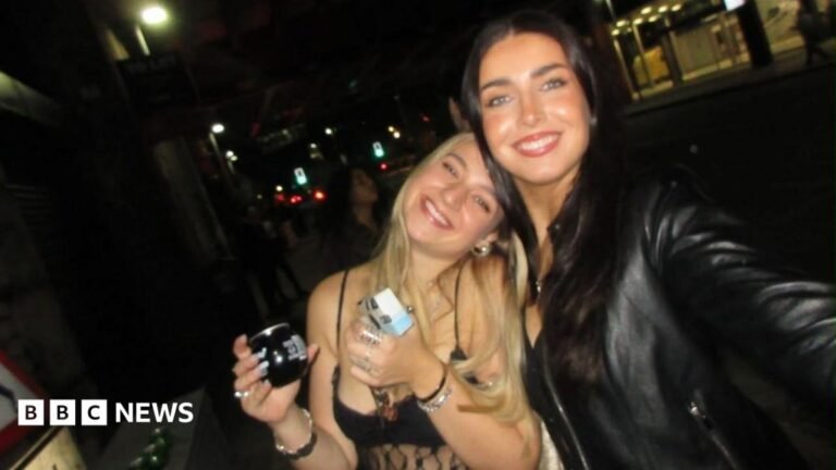 ‘That nagging feeling of dread’: Is ‘hang-xiety’ a real thing? There are two girls in a dark bar smiling at the camera . On the left a blonde girl is wearing a strappy top and holding a drink. She leans into her friend on the left who is holding the camera to take a selfie of the two of them. The girl on the right has long brown hair and is wearing a black leather jacket.