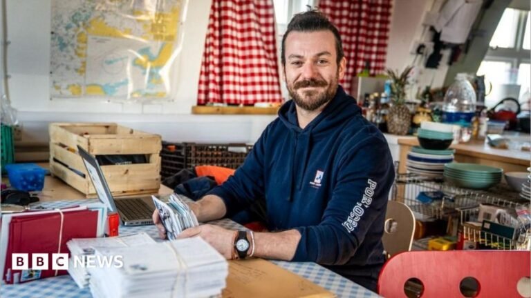 George Clarke sitting at a table with a stack of letters. He is wearing a hoodie with Port Lockroy written on the arm. A kitchen can be seen behind with a sash window.