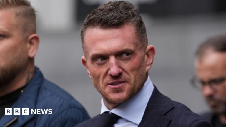 Tommy Robinson stares at the camera as he walks into court, surrounded by a close crowd and security officers. He is wearing a white shirt and black tie. Next to him is a woman with blonde hair and blue eyes, who also stares right at the camera.