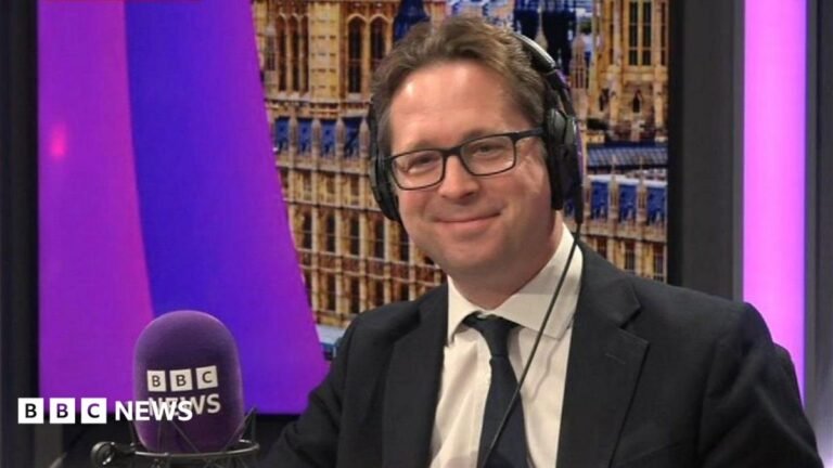 Tories committed to pension triple lock, deputy leader says Andy Burnham is wearing a dark-coloured jacket, a white shirt and glasses. He is stood in front of a red backdrop.