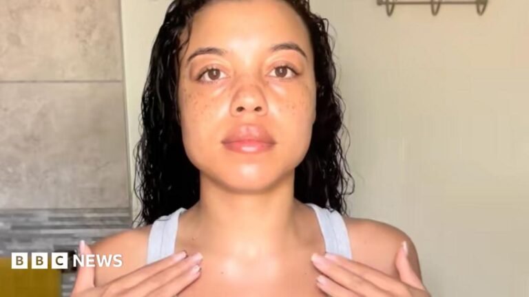 A head and shoulders photo of Abbey Alexandra with long dark curly hair facing the camera wearing a white vest top with her fingertips touching her collarbones