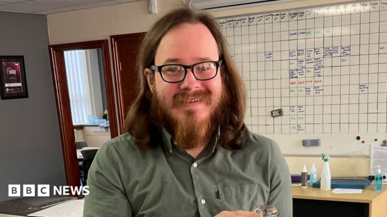 Widdecombe suspect charged with planning acts of terror against Farage A photo of Joshua Kerry, a man with shoulder-length brown hair, a beard and glasses, from the shoulders up. He is smiling at the camera, wearing a khaki shirt, and behind him is a whiteboard.