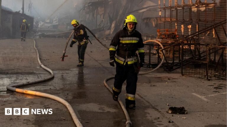 Zelensky condemns ‘vile’ large-scale Russian attacks that killed 24 Three firefighters wearing yellow helmets work at the site of a warehouse that burned out after it was hit during a night of Russian missile attacks in Kyiv. One of the firefighters is seen holding a large hose. The destroyed remains of the warehouse can be seen in the background